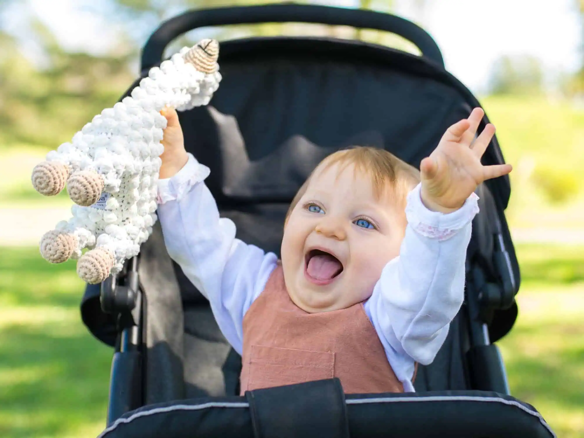 Fröhliches Kleinkind im Kinderwagen hält SindiBaba Lama‑Kuscheltier im Park.