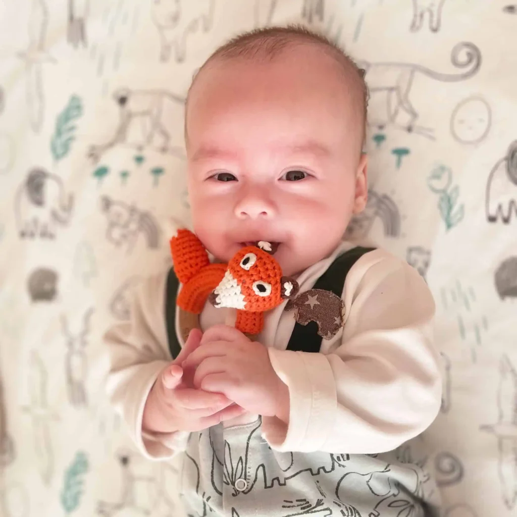 Auf dem Rücken liegendes Baby hält die Ringrassel Fuchs FRED mit beiden Händen und knabbert am Ohr