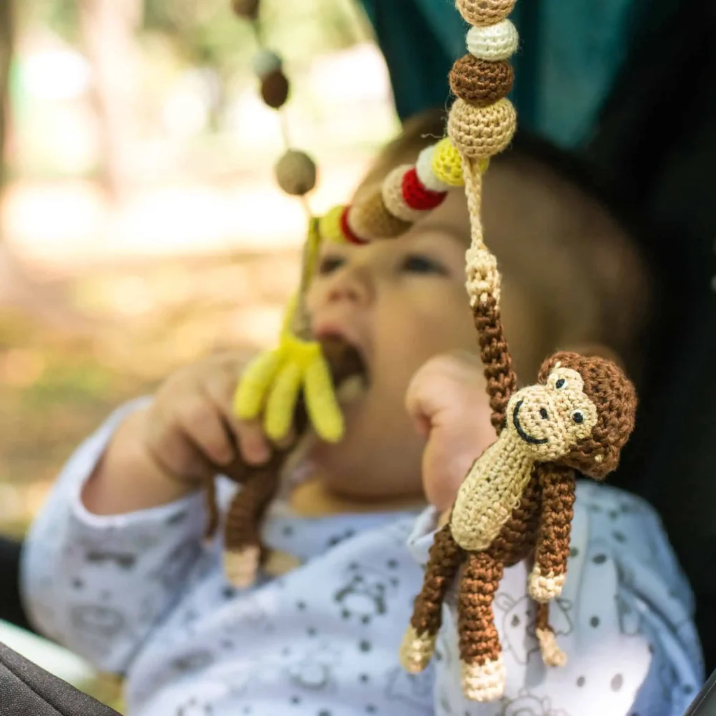 Baby sitzt seitlich im Kinderwagen und beißt auf den Affen der SindiBaba Kinderwagenkette; Park mit Bäumen und Schatten im unscharfen Hintergrund.